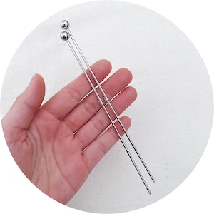 Peut inclure: Deux brochettes en métal argenté avec des extrémités arrondies. Les brochettes sont tenues dans une main.