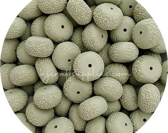 May include: A close-up of a pile of sage green silicone beads with a floral pattern. The beads are oval shaped and have a hole in the centre.