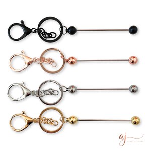 Könnte beinhalten: Vier Schlüsselanhänger mit einem Metallring, einer Metallkette und einer langen Metallstange mit einer Kugel an jedem Ende. Die Schlüsselanhänger sind in Schwarz, Roségold, Silber und Gold erhältlich.