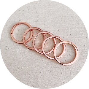 May include: Five rose gold key rings of different sizes, arranged in a circle on a beige fabric background.