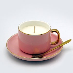 May include: A pink teacup candle with a gold handle and rim, on a matching saucer. A gold spoon rests on the saucer. The candle has white wax and a single wick.
