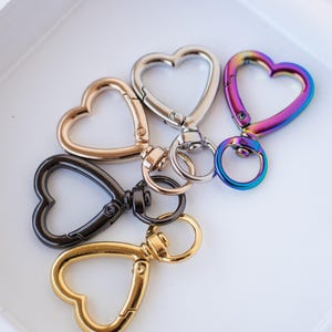 Puede incluir: Una colección de cierres giratorios en forma de corazón en acabados metálicos variados, incluyendo dorado, plateado, negro y un vibrante arcoíris. Estos cierres están diseñados para adjuntar a llaveros, bolsos u otros accesorios.