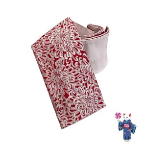 May include: A red and white obi sash with a floral pattern. The sash is folded and tied in a knot. The fabric is brocade with a raised design.