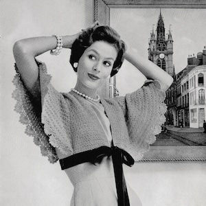 Könnte beinhalten: Eine Frau trägt ein gehäkeltes Bolero mit einer schwarzen Samt-Schärpe, die an der Taille zu einer Schleife gebunden ist. Das Bolero hat einen gewellten Rand und wird über einem weißen Kleid getragen. Die Frau steht vor einem gerahmten Bild einer europäischen Stadt.