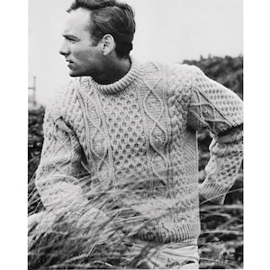 Op de afbeelding: Een zwart-witfoto van een man die een dikke, kabelgebreide trui draagt. De trui heeft een textuurpatroon en is lichtgrijs.