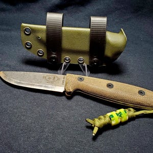May include: A survival knife with a tan handle and a green sheath. The blade has a worn appearance. The handle is secured with black rivets. A green lanyard with yellow and green beads is attached to the handle.