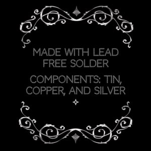 May include: A black background with silver decorative flourishes and text. The text reads "MADE WITH LEAD FREE SOLDER" and "COMPONENTS: TIN, COPPER, AND SILVER". The design is framed by ornate silver scrollwork.