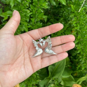 May include: A pair of silver-colored angel wing earrings, each with a heart-shaped center and a star at the top. The earrings are held in an open hand against a backdrop of green foliage.