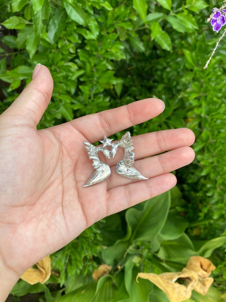 May include: A pair of silver-colored angel wing earrings, each with a heart-shaped center and a star at the top. The earrings are held in an open hand against a backdrop of green foliage.