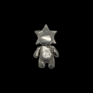 May include: A silver-colored figurine of a humanoid figure with a star-shaped head. The figure has a spiral design on its chest and is set against a black background. The figurine is small and detailed.