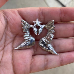 May include: A pair of silver-colored metal angel wing earrings. Each earring features detailed wing designs and a central star-shaped accent. The earrings are displayed against a neutral background.