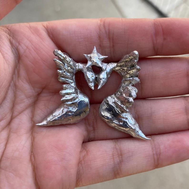 May include: A pair of silver-colored metal angel wing earrings. Each earring features detailed wing designs and a central star-shaped accent. The earrings are displayed against a neutral background.