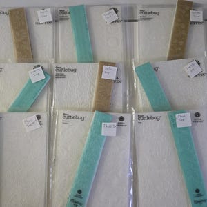May include: A collection of Cuttlebug embossing folders and coordinating paper strips. The folders are in clear plastic packaging, and the paper strips are in shades of teal and tan. Some folders have floral patterns.