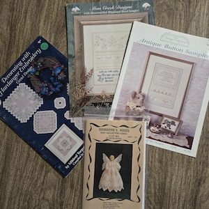 May include: A collection of vintage needlework patterns and samplers, including "Decorating with Hardanger Embroidery" and "Antique Button Sampler." The patterns feature intricate designs and are displayed on a wooden surface.