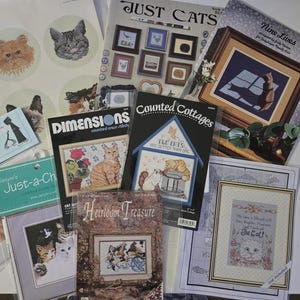 May include: A collection of cat-themed cross-stitch patterns and designs. Includes patterns from Dimensions, Just Cats, and other brands. Features various cat portraits and scenes, with some patterns framed. Ideal for craft enthusiasts.