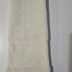 May include: Ivory-colored bath towel, folded lengthwise. The towel features a textured, looped terrycloth body with a woven band near the bottom. The bottom edge is folded over, creating a finished look. The towel is clean and appears unused.