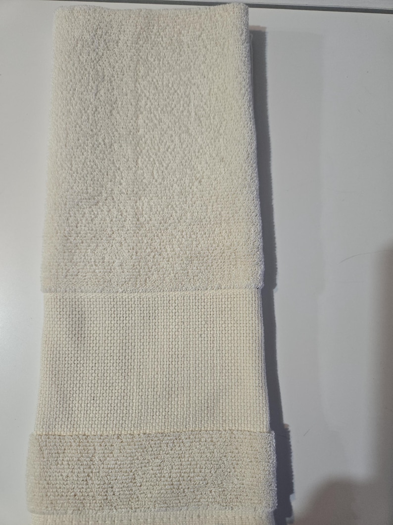 May include: Ivory-colored bath towel, folded lengthwise. The towel features a textured, looped terrycloth body with a woven band near the bottom. The bottom edge is folded over, creating a finished look. The towel is clean and appears unused.