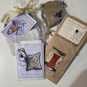May include: A collection of cross-stitch kits and accessories. The image includes a scissor fob, a scissor weight, and a bee-themed scissor case. The scissor fob is labeled "Be Still My Heart". The scissor weight is labeled "giant heart scissor weight".