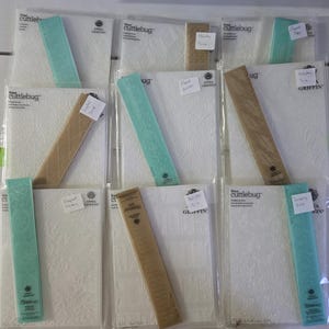 May include: A collection of Cuttlebug embossing folders and decorative strips in clear plastic packaging. The folders are white with embossed patterns, and the strips are various colors, including teal and tan. Some have small white labels.