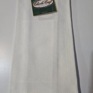 May include: A folded white kitchen towel with a textured weave and a rectangular pocket. A green and gold tag with the text "Charles Craft" is attached to the towel.