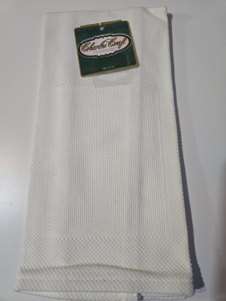 May include: A folded white kitchen towel with a textured weave and a rectangular pocket. A green and gold tag with the text "Charles Craft" is attached to the towel.