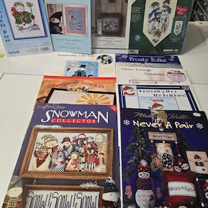 May include: A collection of snowman-themed cross-stitch kits and patterns. The kits feature various designs, including snowmen with hats and scarves, and banners with holiday themes. The image includes the text "Snow Fun" and "Snowman Collector".