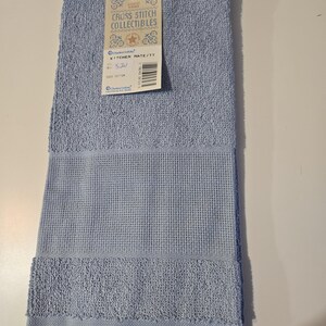 May include: A light blue kitchen towel with a textured waffle weave and fringe detail. The towel has a tag that reads "Cross Stitch Collectibles" and "Kitchen Mate/TT". Made of 100% cotton.