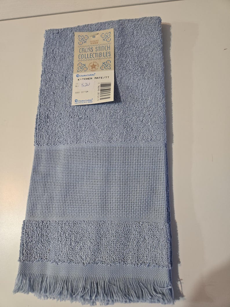May include: A light blue kitchen towel with a textured waffle weave and fringe detail. The towel has a tag that reads "Cross Stitch Collectibles" and "Kitchen Mate/TT". Made of 100% cotton.