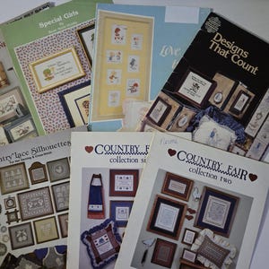 May include: A collection of seven softcover cross-stitch pattern booklets featuring various designs. Titles include Through with the Sun, Special Girls, Designs That Count, Country Lace Silhouettes, and Country Fair collections one and two.