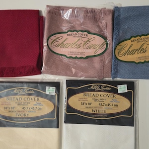 May include: An assortment of bread covers and napkins in various colors. The image includes a red napkin with fringe, pink and blue bread covers in plastic packaging, and ivory and white bread covers in packaging. The bread covers are 18 x 18 inches.