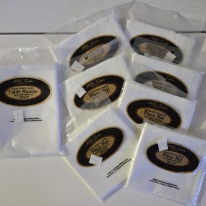 May include: A collection of white cross-stitch fabric items, including a table runner and place mats, packaged in clear plastic. Each item has a black and gold oval label with text. The table runner is labeled "Table Runner". The place mats are labeled "Place Mat".