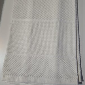 May include: A folded white kitchen towel with a textured diamond pattern on the top and bottom sections, and a horizontal band of a different texture in the middle. The towel is made of a soft, absorbent material.