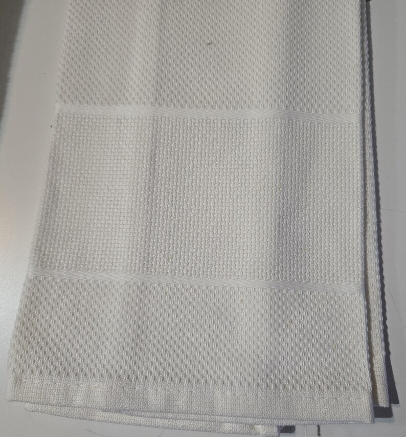 May include: A folded white kitchen towel with a textured diamond pattern on the top and bottom sections, and a horizontal band of a different texture in the middle. The towel is made of a soft, absorbent material.