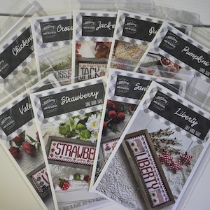 May include: A collection of cross-stitch kits in clear plastic bags. The kits feature various designs, including "Chickens," "Cross," "Jack," "Pumpkins," "Strawberry," "Valentine," "Santa," and "Liberty." Each kit has a black and white gingham border.
