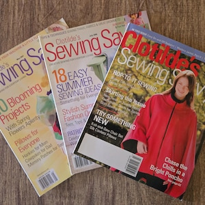 May include: Three issues of "Clotilde's Sewing Savvy" magazine are displayed on a wooden surface. The magazines feature articles on sewing projects, summer ideas, and fashion. The covers showcase colorful designs and text.