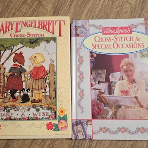 May include: Two cross-stitch pattern books. The book on the left is titled "Mary Engelbreit Cross-Stitch Make a Wish". The book on the right is titled "Alma Lynne's Cross-Stitch for Special Occasions".