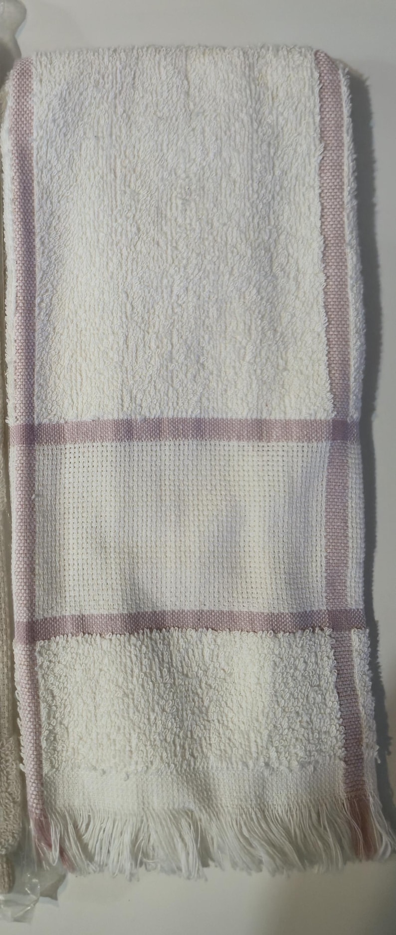 May include: White bath towel with a textured surface and a woven section with a lavender stripe. The towel has a fringe at the bottom edge. A simple and elegant design for the bathroom.