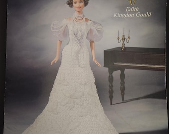 May include: A porcelain doll wearing a white lace dress and a pearl necklace. The doll is part of The Gilded Age Collection and is named Edith Kingdon Gould. The doll is standing in front of a piano with a candelabra on top.