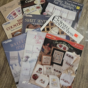 May include: A collection of cross-stitch pattern booklets with titles like "Tender Touches for Fingertips," "Sweet Sentiments," and "Life on the Farm." The covers feature images of embroidered designs, farm scenes, and floral motifs. The booklets are arranged on a wooden surface.