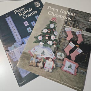 May include: Two Green Apple Co. craft pattern booklets. The left booklet is titled Peter Rabbit Counts, Book 517, featuring a blue counted cross-stitch wall hanging. The right booklet is titled Peter Rabbit Christmas, Book 516, showing a decorated tree.