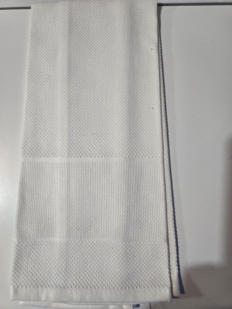 May include: A folded white kitchen towel with a textured diamond pattern. The towel has a rectangular shape with a horizontal band of a different texture. The towel is made of a soft, absorbent material.