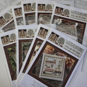 May include: A collection of cross-stitch patterns in clear plastic sleeves. The patterns feature images of houses, trees, and farm scenes. The text "Little House Needleworks" and "Fall on the Farm" are visible on the packaging.