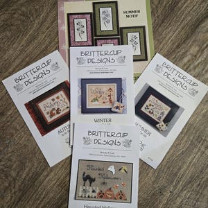 May include: A collection of cross-stitch pattern booklets from Brittercup Designs, featuring seasonal themes. The booklets showcase designs for summer, autumn, winter, and a haunted Halloween. The covers display framed cross-stitch examples with floral and holiday motifs.