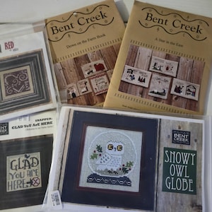 May include: Assortment of cross-stitch patterns and booklets. Includes framed cross-stitch designs with heart and owl motifs, and booklets titled "Down on the Farm Book" and "A Star in the East." The "Snowy Owl Globe" design is also visible.