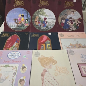 May include: A collection of Precious Moments books and cross-stitch patterns. The books have illustrations of children and biblical scenes. The cross-stitch patterns feature the Precious Moments girl. The color palette includes burgundy, blue, and pink.