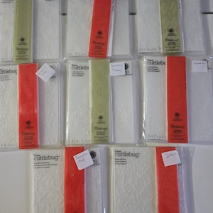 May include: A collection of Cricut Cuttlebug embossing folders in clear plastic packaging. The folders feature various embossed designs, including floral and geometric patterns. Some packages have red or green accents.