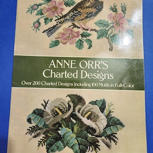 May include: Book cover featuring a cross-stitch design of a bird perched on a branch with pink flowers. The title of the book is "ANNE ORR'S Charted Designs" and the subtitle is "Over 200 Charted Designs including 100 Motifs in Full-Color".
