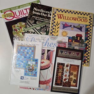 May include: A collection of quilting books and magazines. Titles include "Quilt News," "Needlecraft from Our Best," and "Willowood." The image features various quilt patterns and designs, including fish, hearts, and fall-themed motifs. The books are in various sizes and colors.