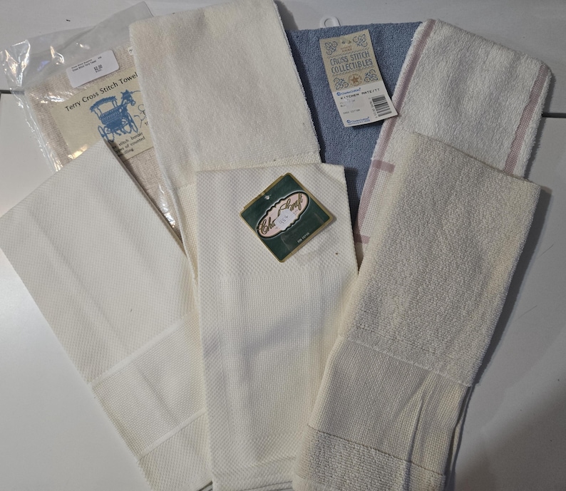 May include: A collection of various white and light-colored kitchen towels. Some towels have textured patterns, while others are smooth. One towel has a tag that reads "Terry Cross Stitch Towel" with an image of a horse-drawn carriage. Another tag reads "Crown Stitch Collectibles".