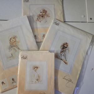 May include: Several cross-stitch kits featuring portraits of women in vintage clothing. The kits are packaged in clear plastic and beige paper, with the "Lanarte" logo visible. The artwork includes soft colors and delicate details.